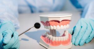 A dental doctor wearing blue gloves and a mask holds a dental model of the upper and lower jaws and a dental mirror.